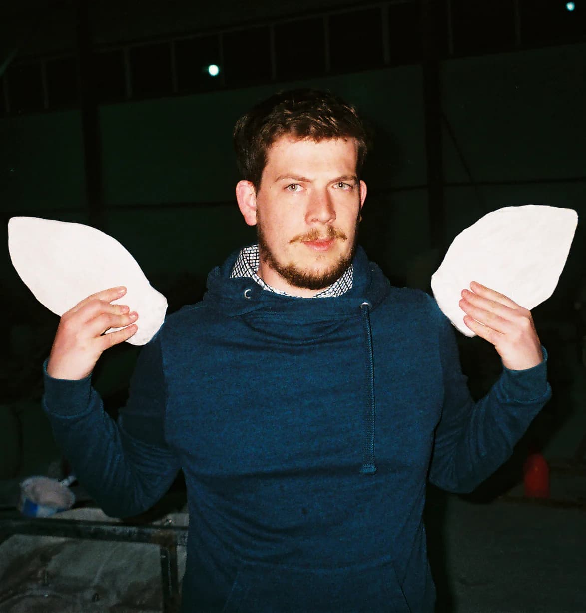 Alberto stands in a darkened workshop space, wearing a blue hoodie over a collared shirt, holding up two stegosaurus scales at shoulder height — one in each hand — and looking directly at the camera with a composed expression.
