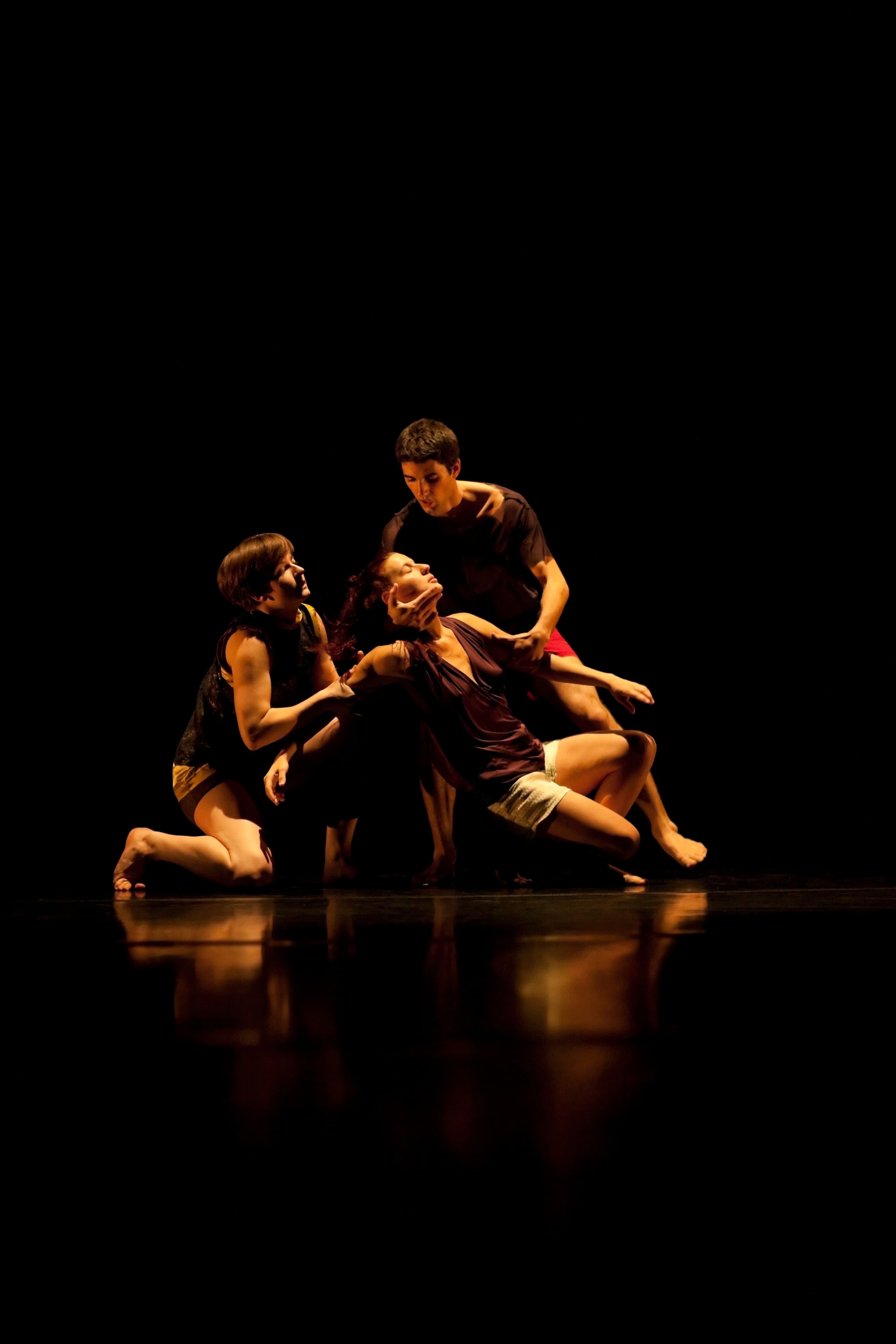 Three performers on stage creating the image of la pieta with the dying character female and a male and female character holding her, creating a triangle in height, washed by warm golden light, all wearing shorts
