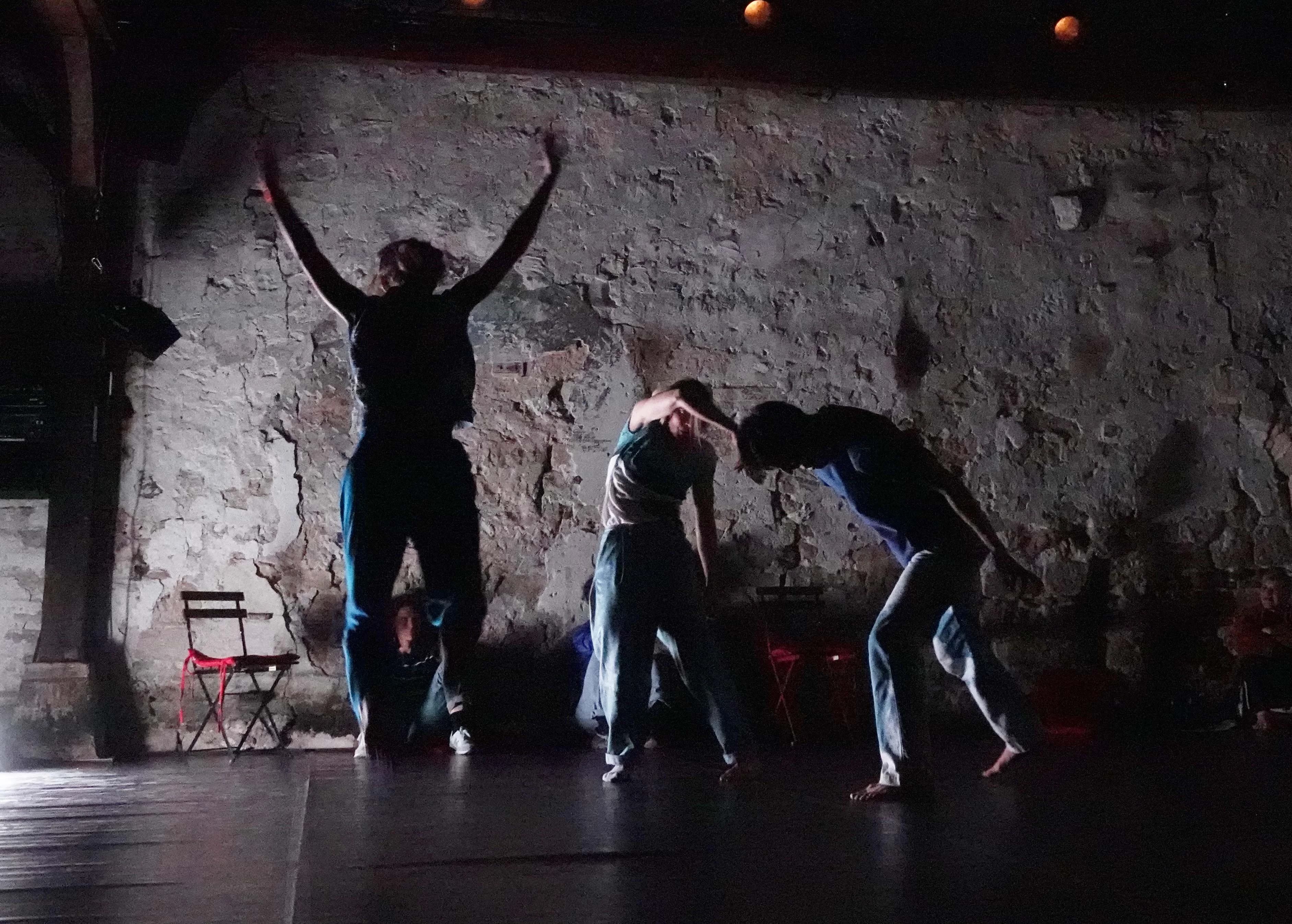 Three performers on stage against a weathered brick and concrete wall in a darkened performance space. The figure on the left has arms raised triumphantly, while the center and right performers are captured mid-movement in crouched or bent positions.