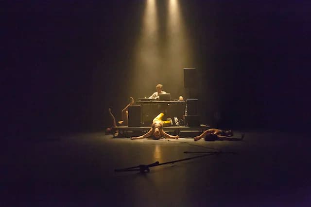 Stage with a platform where a musician, Alberto, playing with their laptop, with three people dead falling from the platform, lit by two epic golden cenital lights