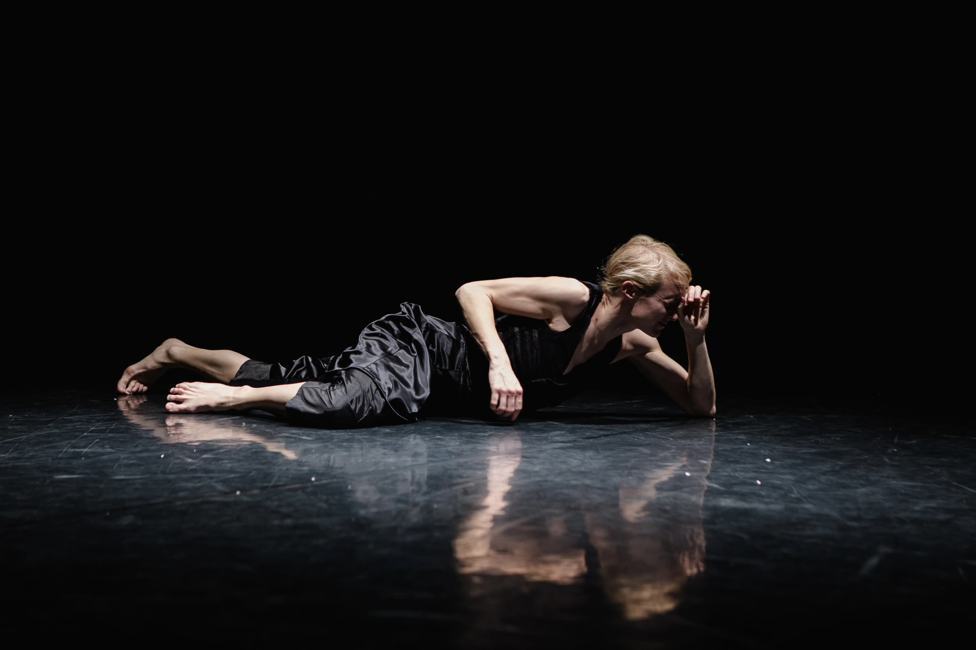 A dancer dressed in black silky jumpsuit with blonde hair lies horizontally across a dark, reflective stage floor. Their left hand against their face creating a peeping whole they are looking through.