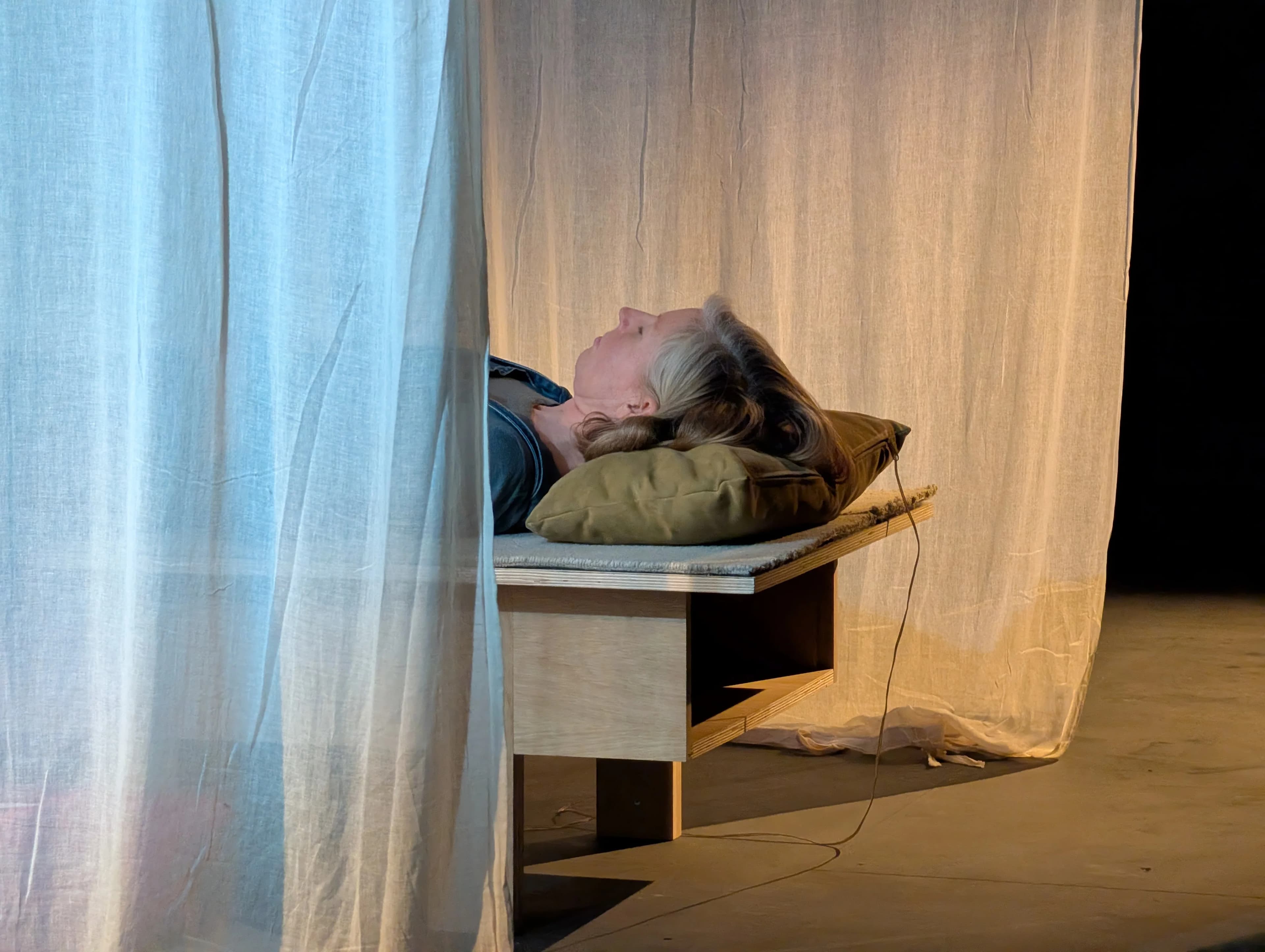 A person lies on their back on a minimalist wooden bench with a green cushion pillow, eyes closed in a restful, contemplative pose.