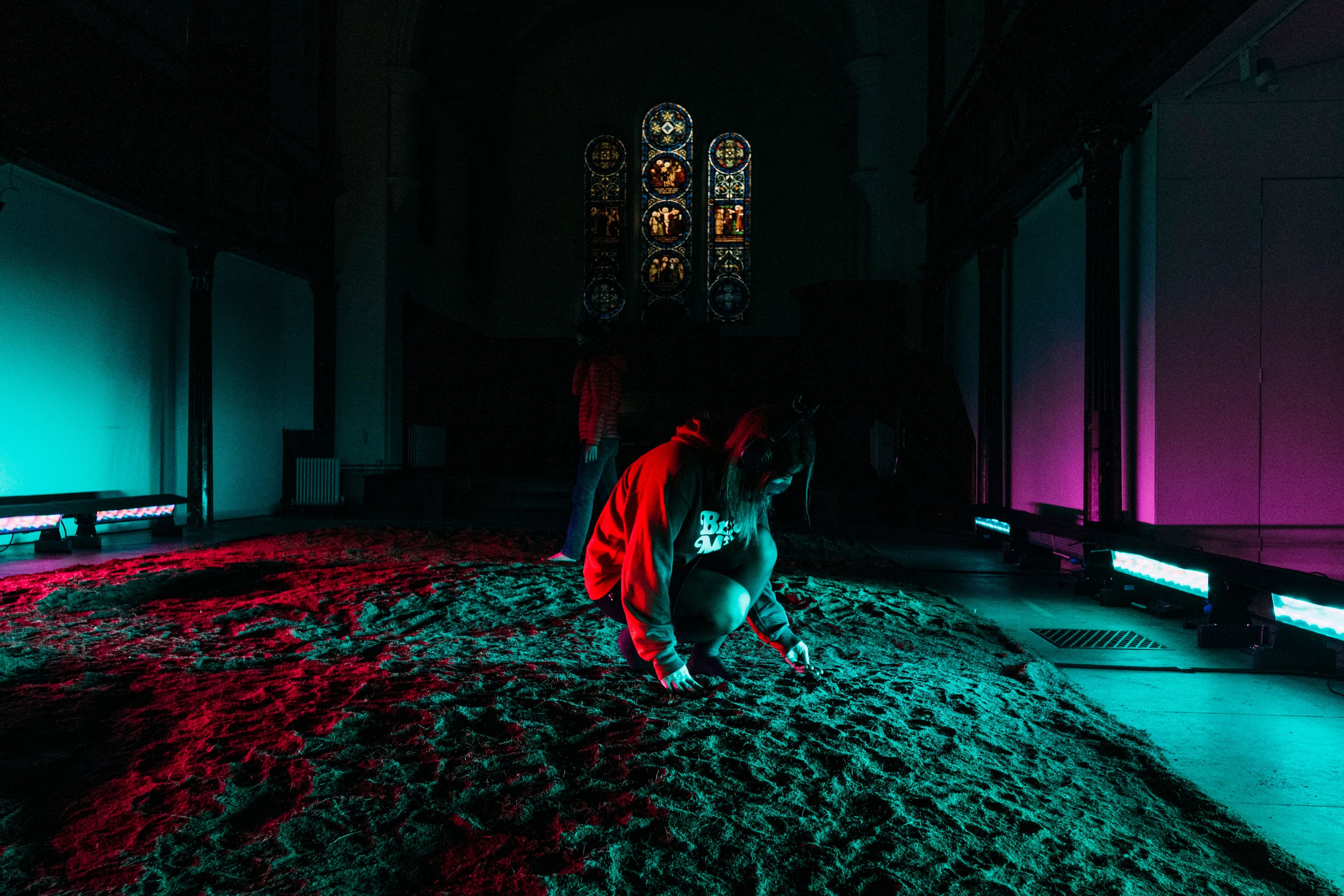 Installation consisting of a surface of soil on the ground lit with colourful lights knee height inside a church with a person barefoot crouched touching the soil and wearing headphones