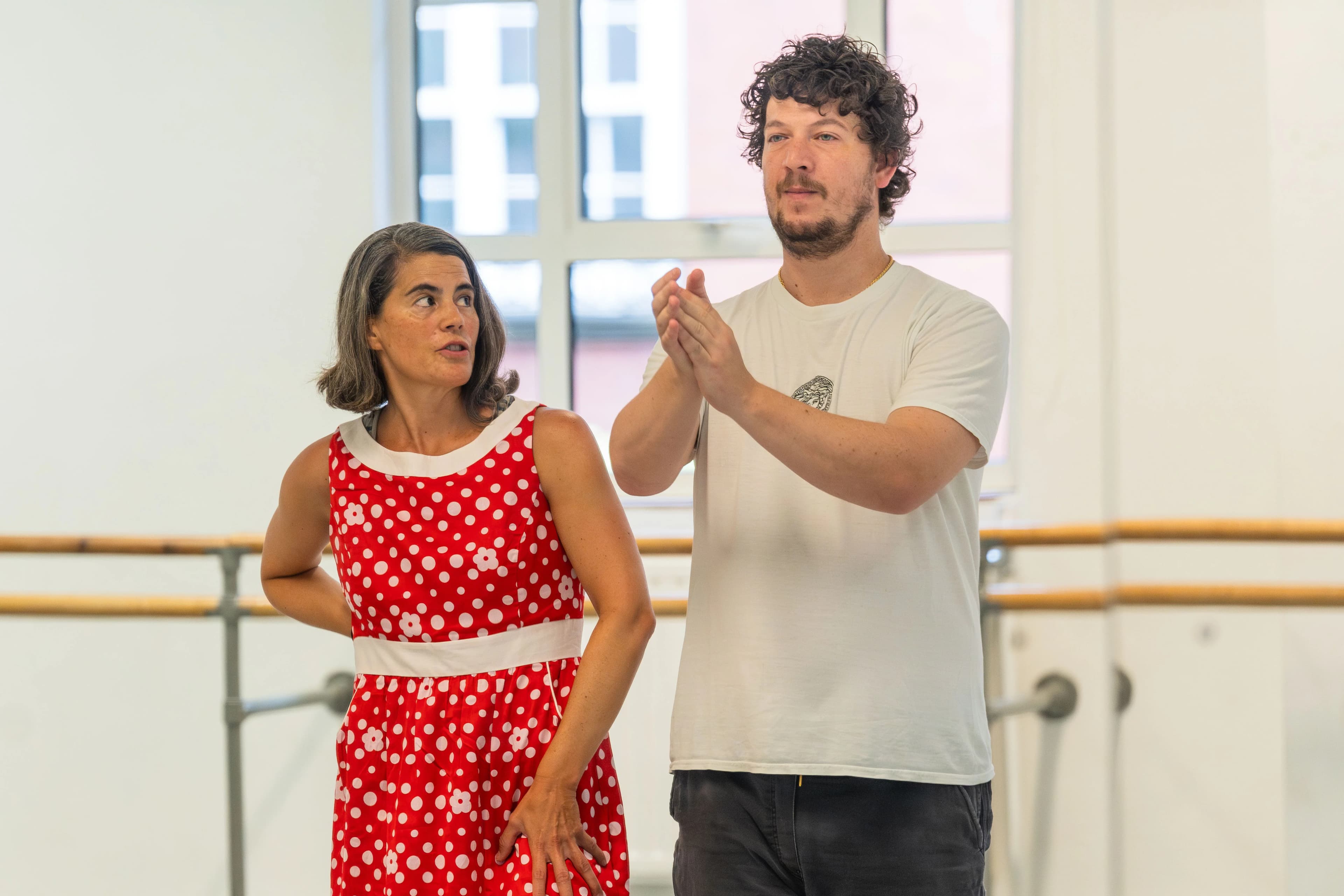 Eva wearing a red and white polka dot dress and Alberto in a white t-shirt stand together in a bright dance studio. Eva looks pensively to the side while Alberto claps their hands.