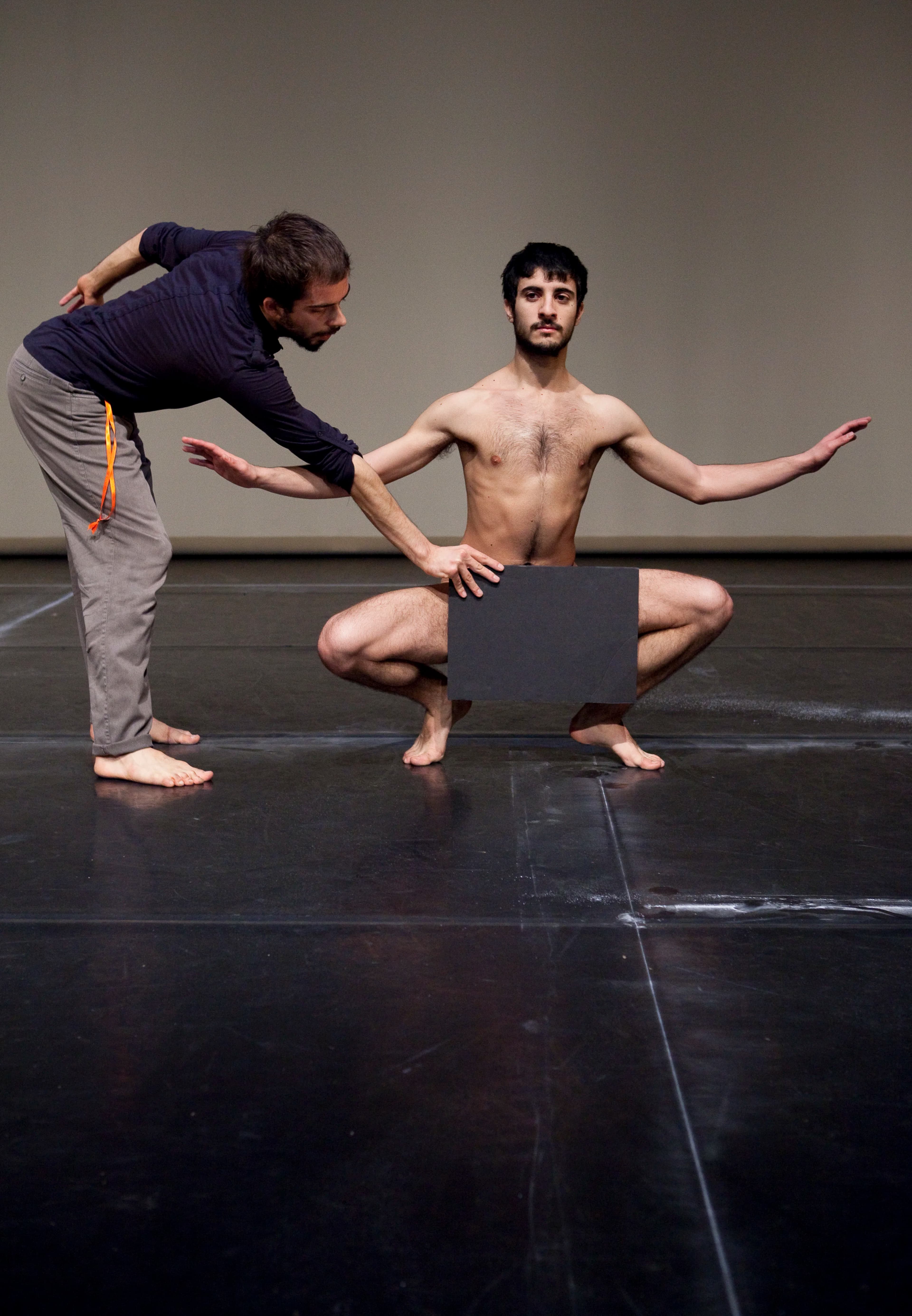 Two men, one naked crouching and one dressed covering the other man's genital area with a black panel