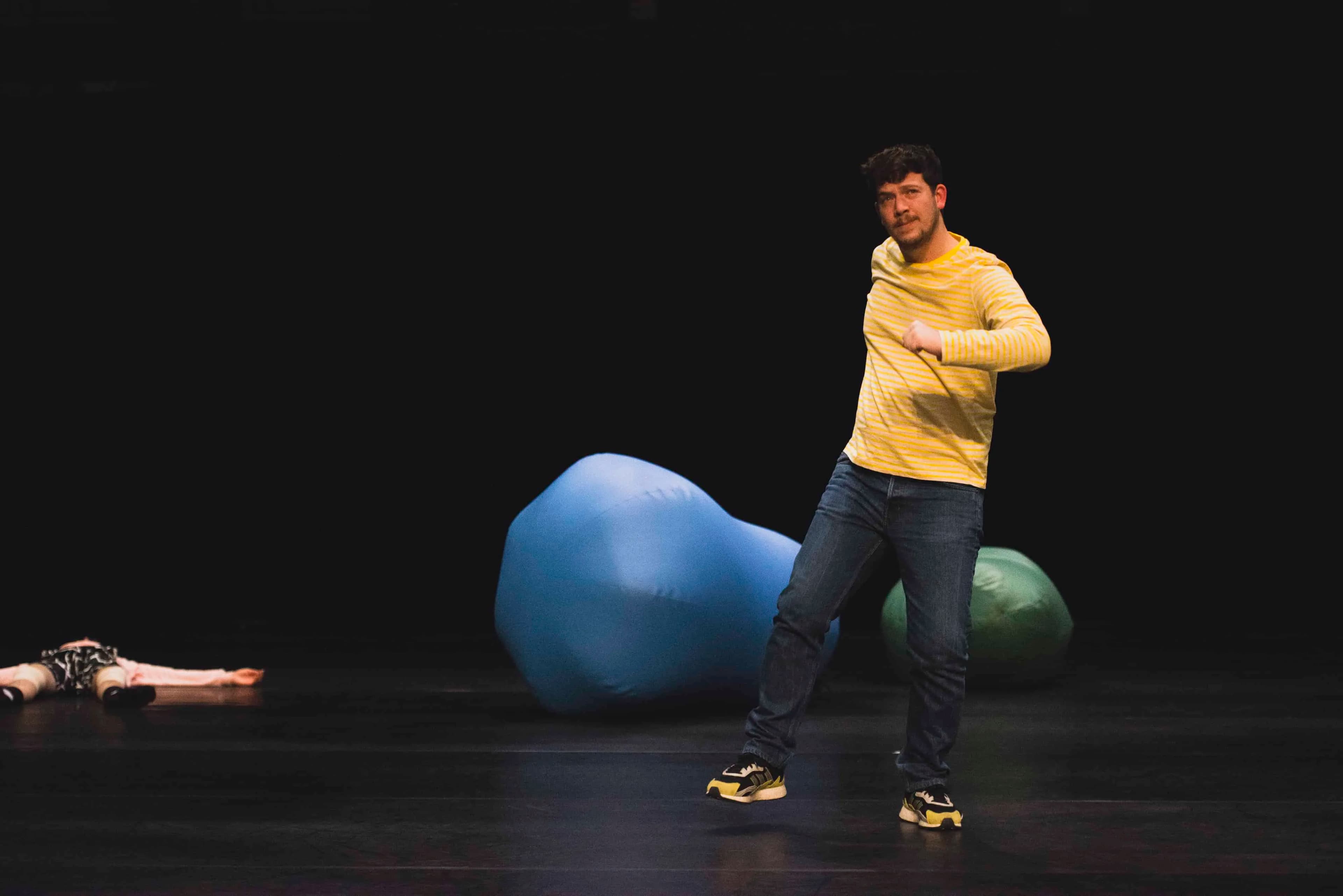 Black stage with Alberto in the foreground mid movement wearing yellow t-shirt and shoes, with a person lying dead and large sacks in the background