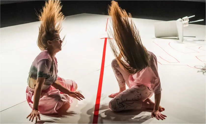 Two women on a white stage dancing with their hair moving around, with red markings on the floor and a white chair lying on the floor