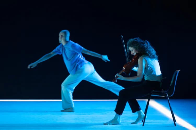 Jesús Rubio Gamo, a male dancer, is captured mid-lunge with extended arms on stage, while Luz Prado, a violinist, sits on a black chair playing her violin beside him. Both are illuminated by vibrant blue stage lighting against a dark background, creating an intimate moment that visually expresses the dialogue between dance and music.
